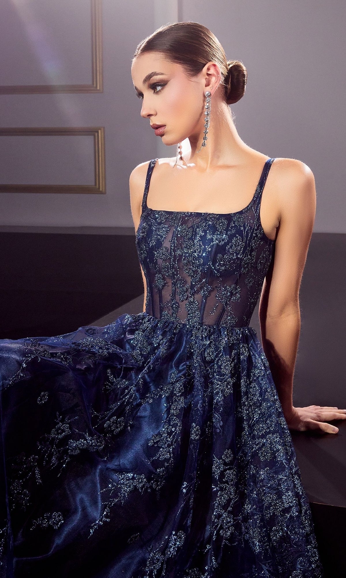 A woman in the Long A-Line Glitter-Print Prom Dress J840 sits on a dark surface, wearing long earrings and looking to the side with her hair styled in a sleek bun.